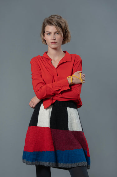 Woman wearing red top and wool skirt. Wool skirt has colour block pattern made from blue, burgundy, red, black and white colours.